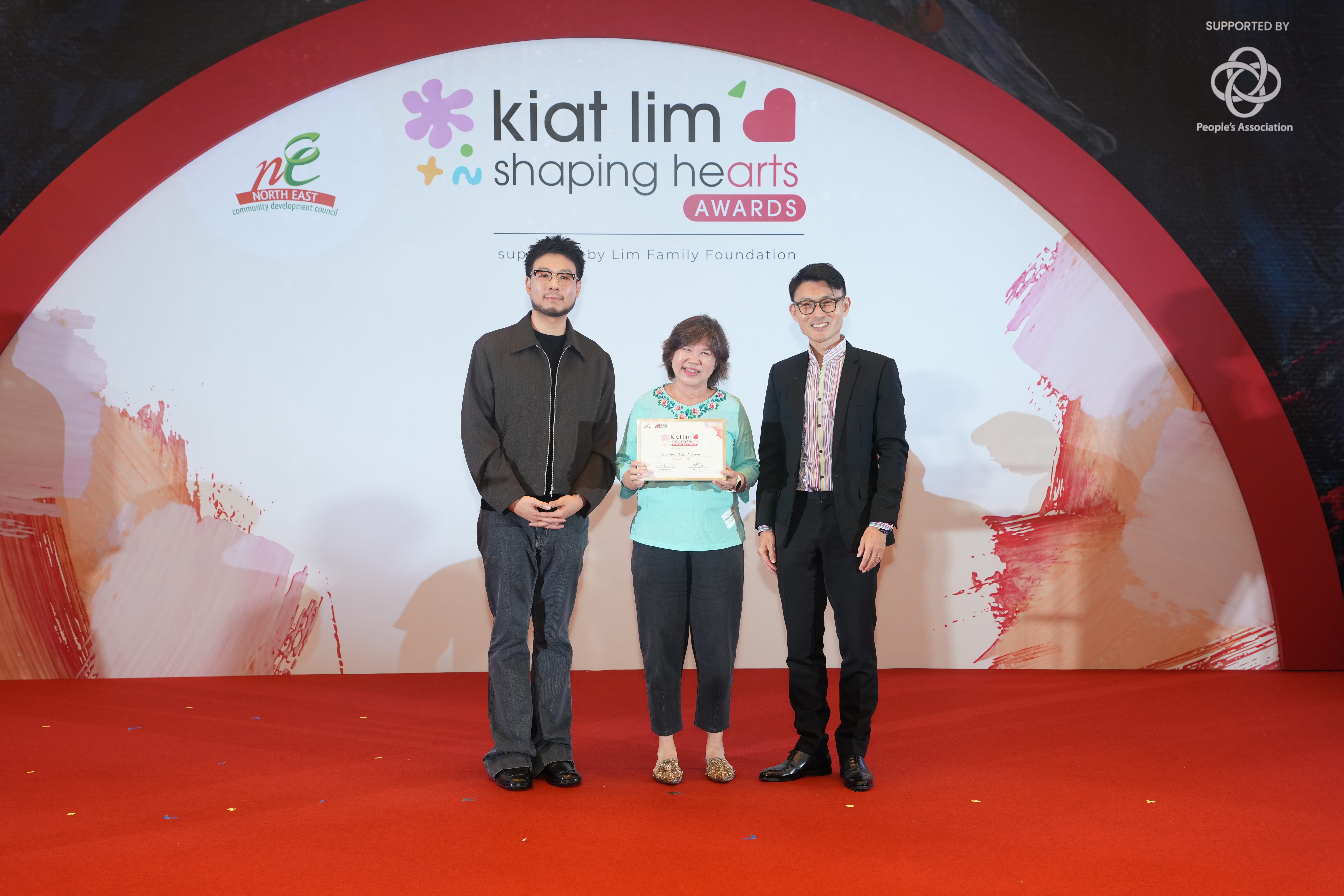 Award recipient standing on stage with the presenter, holding their certificate and posing for a group photo during the award ceremony, with a backdrop reading ‘Kiat Lim-Shaping Hearts Awards'.